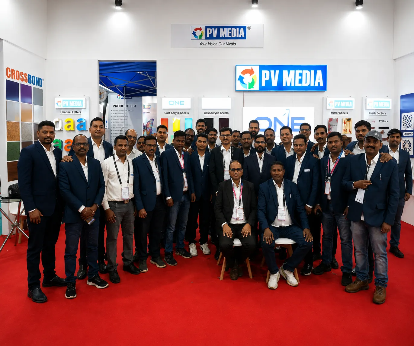 Large group of PV Media staff posing indoors at a trade show booth displaying Channel Letters, Cast Acrylic Sheets, and Mirror Sheets.