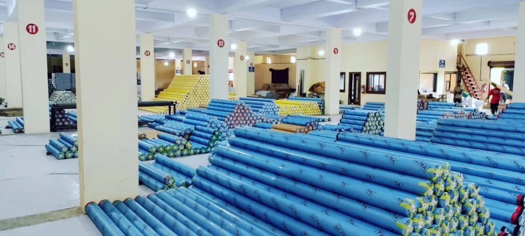 Stacks of blue 'ULTRA' wrapped material rolls fill a warehouse with numbered pillars and workers visible in the background.
