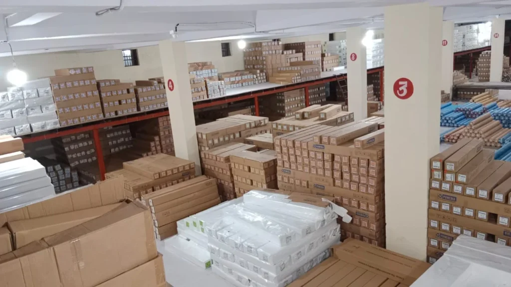 Bright warehouse interior displays stacks of boxes and blue-wrapped rolls on shelves and the floor.